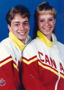 A young man and woman posing in matching team jackets for Canada. The jacket has a yellow collar with white inside, a white upper portion with the words in Canada in red followed by red lower part with a yellow diagonal stripe.