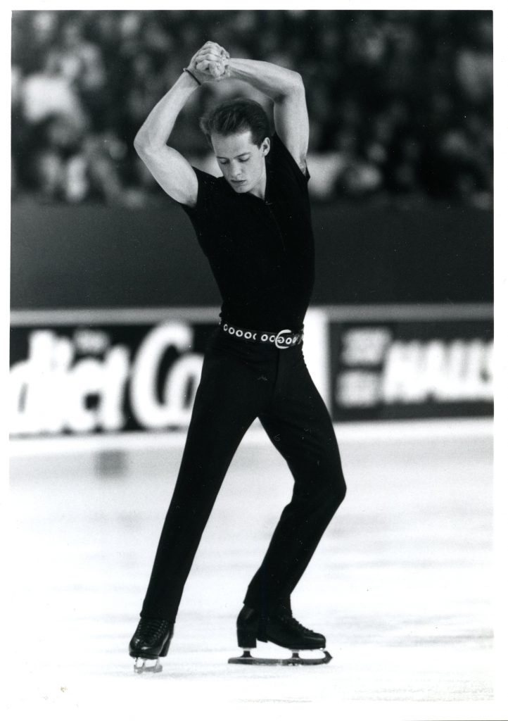 Skater Kurt Browning in a pose with his arms over his head and one leg bent and the other stretched. His outfit is a black fitted short-sleeved top and pant legs. He wears a belt with large circular metal grommets.