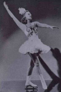 Sonja Henie in a fancy skating costume posing for the camera with both arms extended, one up behind her head and the other extended in front.